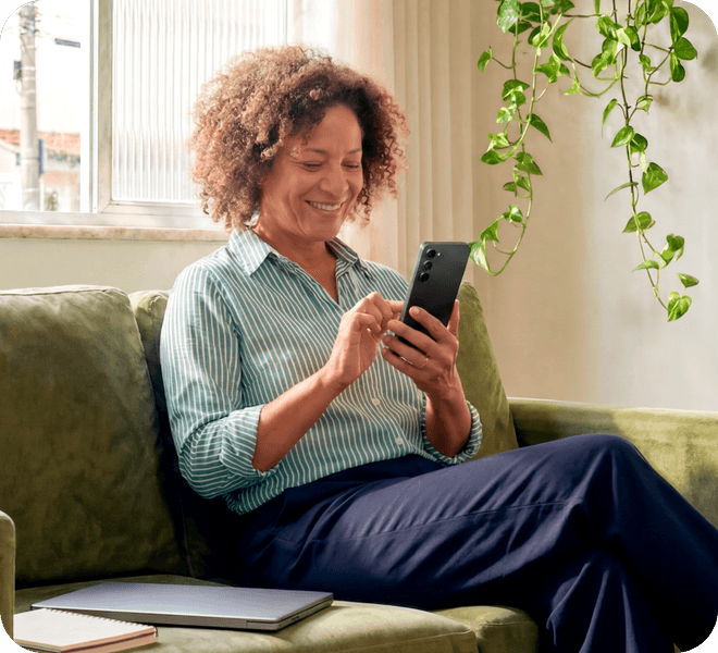 Empreendedora no sofá de casa sorrindo e observando a tela do celular nas mãos