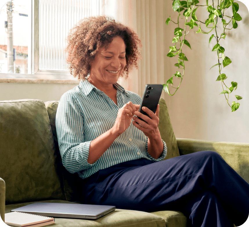 Empreendedora no sofá de casa sorrindo e observando a tela do celular nas mãos
