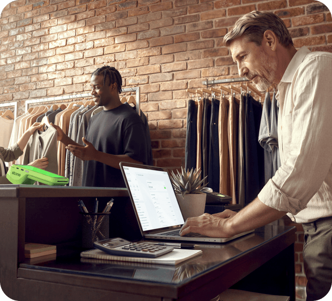 Empreendedor maduro em loja de roupas analisando vendas no notebook, com funcionário atendendo cliente ao fundo e maquininha verde Stone na mesa