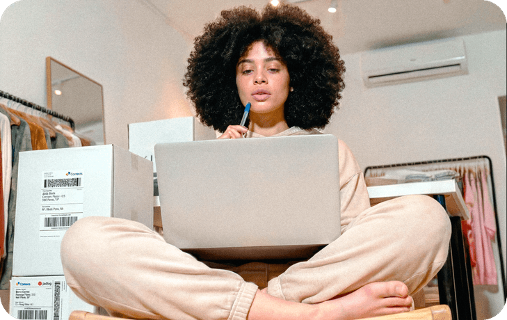 Empreendedora de cabelo afro em loja de roupas sentada com notebook, com caixas dos Correios ao lado para envios