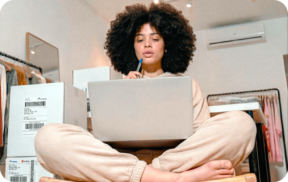 Empreendedora de cabelo afro em loja de roupas sentada com notebook, com caixas dos Correios ao lado para envios