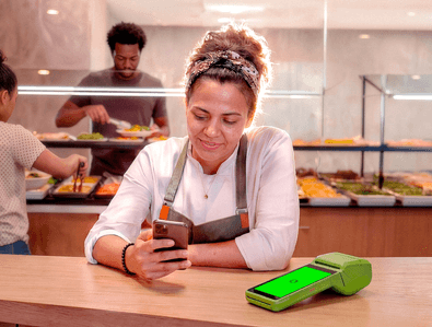 Atendente de restaurante self-service mexendo no celular ao lado da maquininha Stone