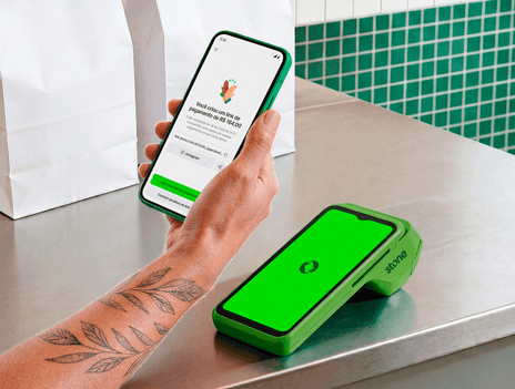 Celular com tela do app Stone confirmando a criação de um link de pagamento de R$ 100,00, ao lado da maquininha verde