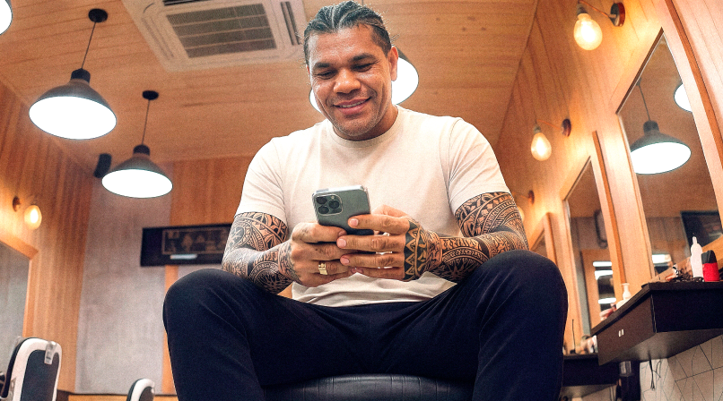 Barbeiro tatuado sentado em barbearia sorrindo ao consultar o celular