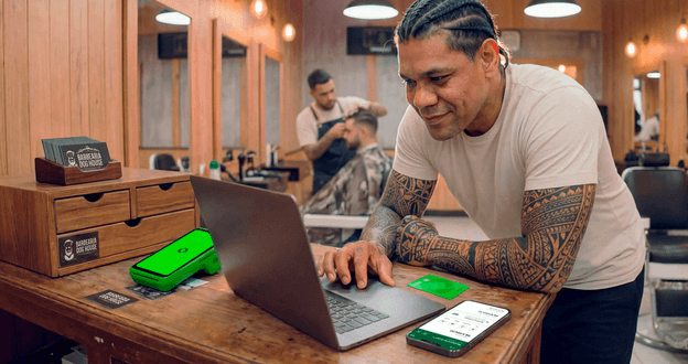 Barbeiro tatuado em barbearia analisando vendas no notebook, com maquininha Stone e cartões da loja no balcão
