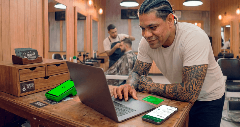 Barbeiro tatuado em barbearia analisando vendas no notebook, com maquininha Stone e cartões da loja no balcão