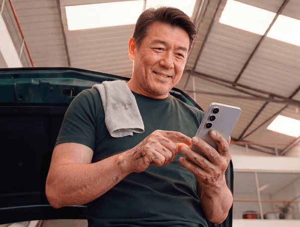 Mecânico em oficina sorrindo ao usar o celular, com pano apoiado no ombro