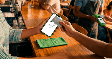 Cliente em bar segurando o celular para pagar com cartão pela Conta Stone, com atendente ao fundo segurando a maquininha verde