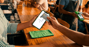 Cliente em bar segurando o celular para pagar com cartão pela Conta Stone, com atendente ao fundo segurando a maquininha verde