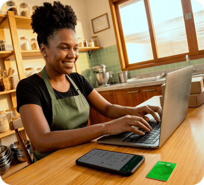 Pessoa empreendedora sorrindo enquanto usa um notebook na cozinha do seu negócio, com um celular e um cartão Stone sobre a mesa.