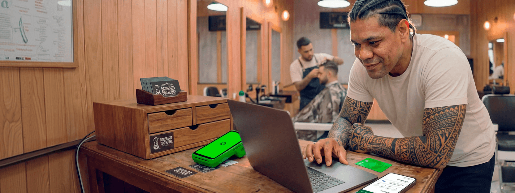 Barbeiro tatuado em barbearia analisando vendas no notebook, com maquininha Stone e cartões da loja sobre a bancada