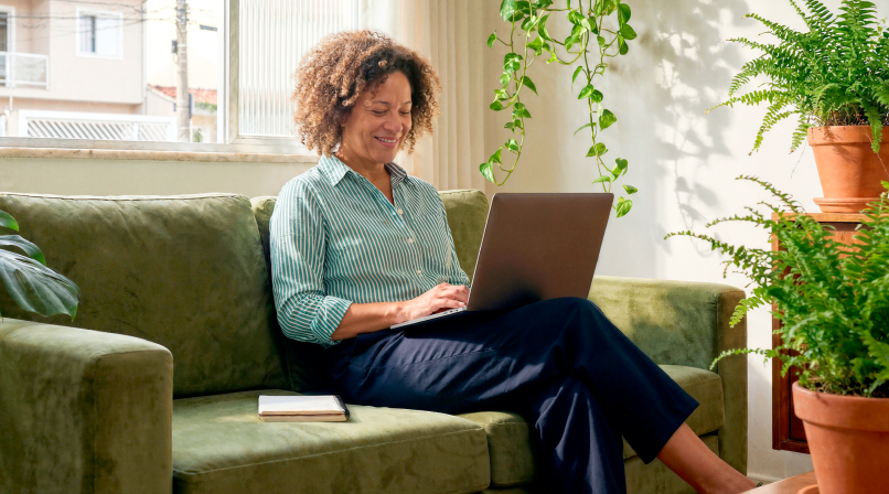 Mulher madura sorrindo em casa sentada no sofá usando o notebook