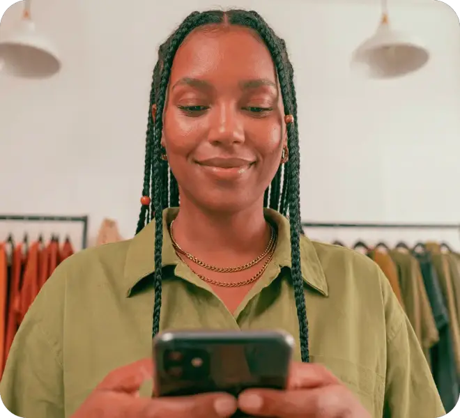 A empreendedora em sua loja de roupas, com linhas de costuras atrás está segurando um celular para enviar o link de pagamento aos seus clientes através do Messenger do Facebook, Whatsapp e Instagram.