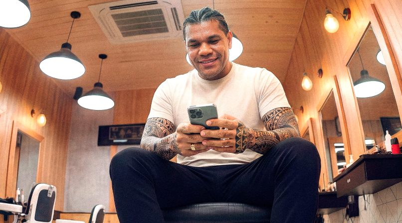 Barbeiro tatuado em barbearia sorrindo enquanto consulta o celular