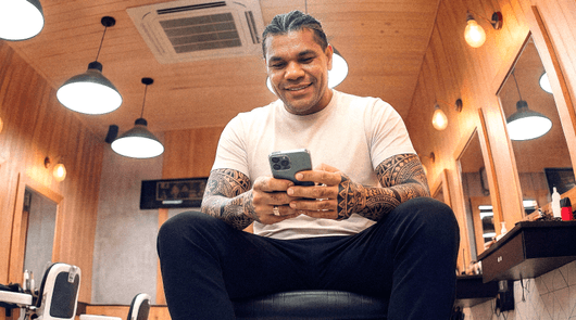 Barbeiro tatuado em barbearia sorrindo enquanto consulta o celular