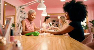 Manicure pintando as unhas de uma cliente em estúdio rosa com a maquininha Stone na mesa
