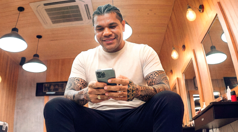 Barbeiro tatuado sentado em barbearia sorrindo enquanto usa o celular