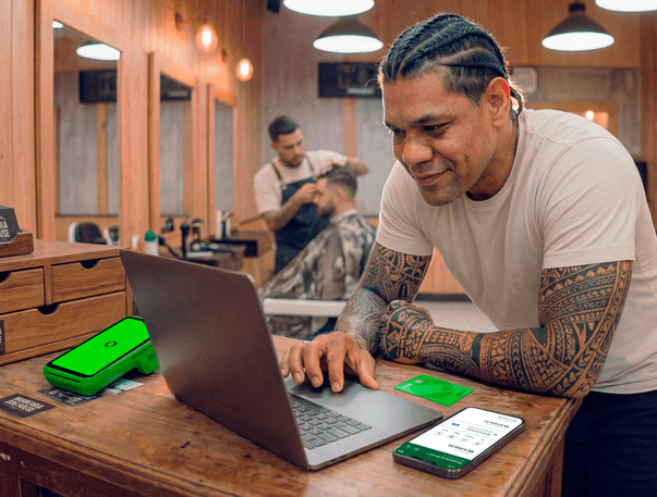 Barbeiro tatuado em barbearia analisando vendas no notebook, com maquininha Stone e cartões da loja sobre a bancada