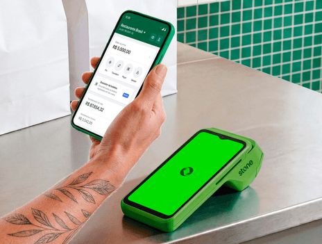 Atendente em restaurante segurando o celular com app Stone aberto ao lado da maquininha Stone com logo na tela