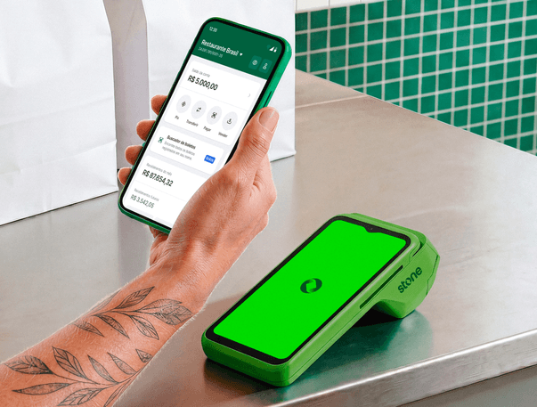 Atendente em restaurante segurando o celular com app Stone aberto ao lado da maquininha Stone com logo na tela