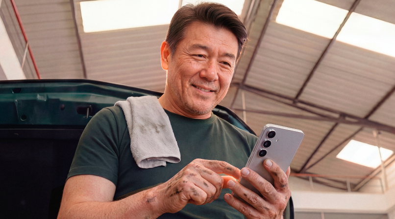 Mecânico em oficina sorrindo ao consultar o celular, com pano apoiado no ombro