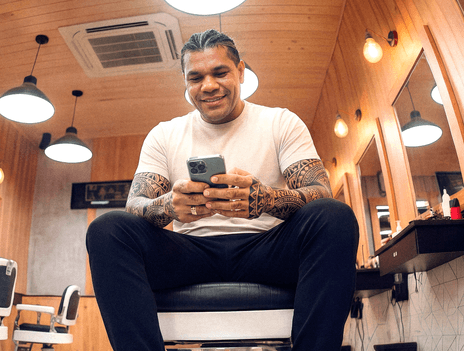 Barbeiro tatuado sentado em cadeira de barbearia consultando o celular