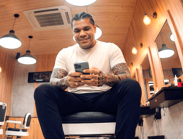 Barbeiro tatuado sentado em cadeira de barbearia consultando o celular