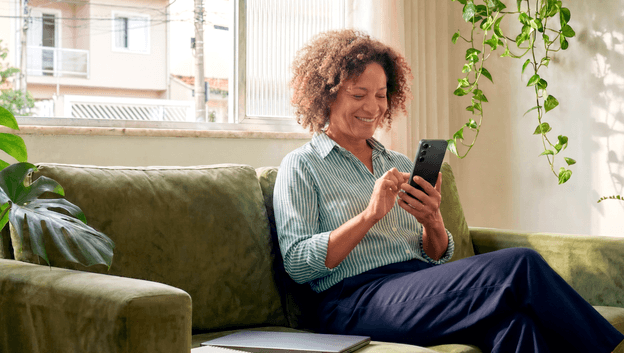 Mulher madura sorridente sentada no sofá de casa consultando o celular