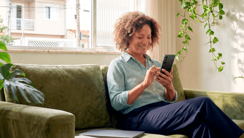 Mulher madura sorridente sentada no sofá de casa consultando o celular