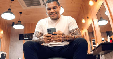 Barbeiro tatuado sentado em barbearia sorrindo enquanto consulta o celular