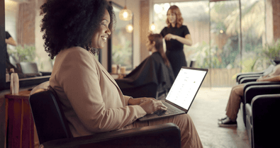 Empreendedora sorridente em salão de beleza usando o notebook enquanto outra cliente é atendida ao fundo