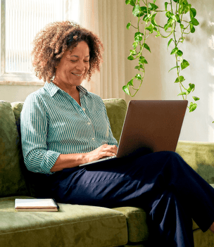 Mulher madura sorridente sentada no sofá de casa usando o notebook