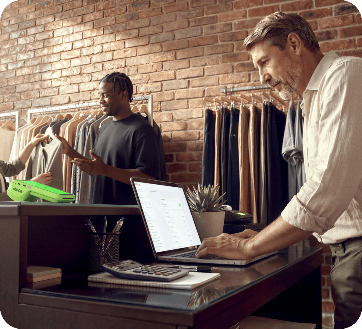 Empreendedor maduro em loja de roupas analisando vendas no notebook, com funcionário atendendo cliente ao fundo e maquininha verde Stone na mesa