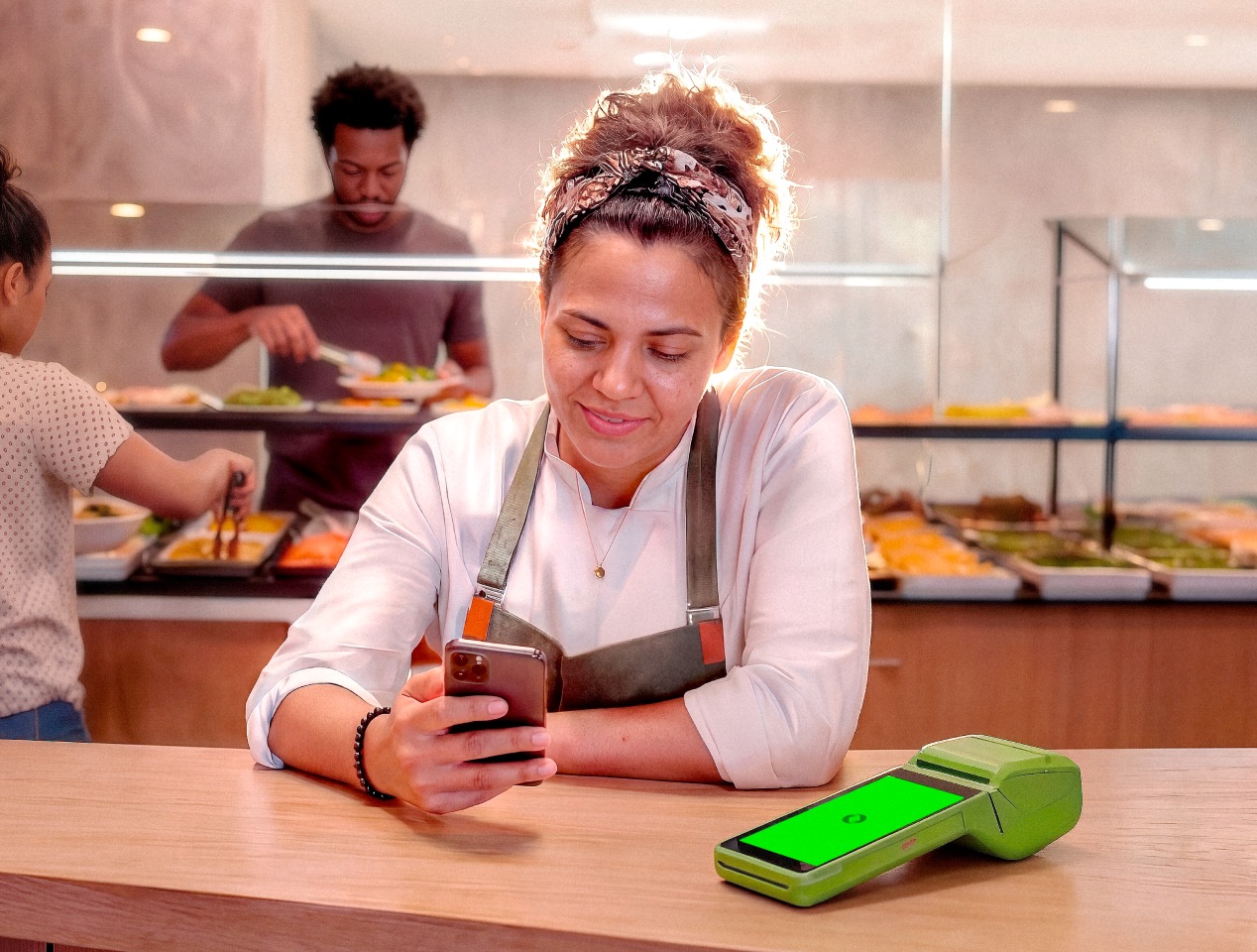 Atendente de restaurante self-service mexendo no celular ao lado da maquininha Stone