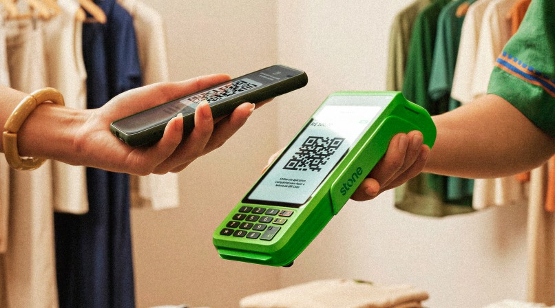 Cliente em loja de roupas pagando por QR Code Pix com o celular escaneando o código na maquininha Stone