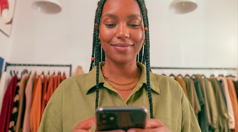 Mulher sorrindo, olhando para a tela do celular em suas mãos e com uma arara de roupas ao fundo