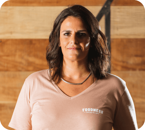 Re Cruz, diretora executiva e criativa da Foodness, vestindo uma camiseta rosé claro da marca, em frente a um fundo de madeira.