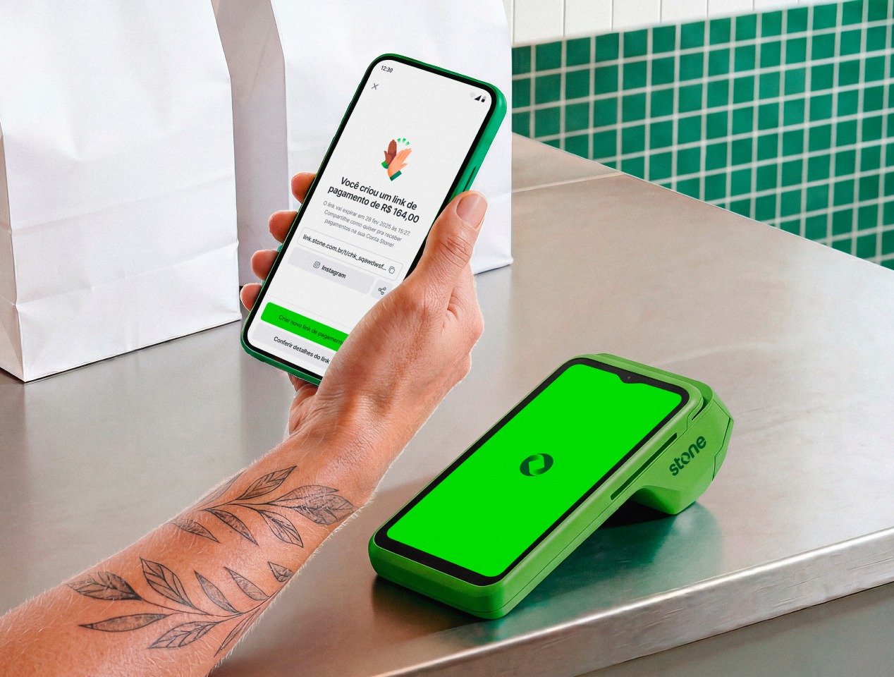 Celular com tela do app Stone confirmando a criação de um link de pagamento de R$ 100,00, ao lado da maquininha verde