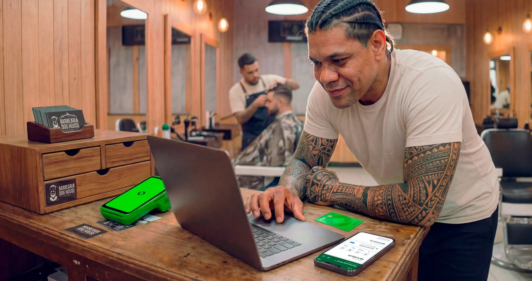 Barbeiro tatuado em barbearia analisando vendas no notebook, com maquininha Stone e cartões da loja no balcão