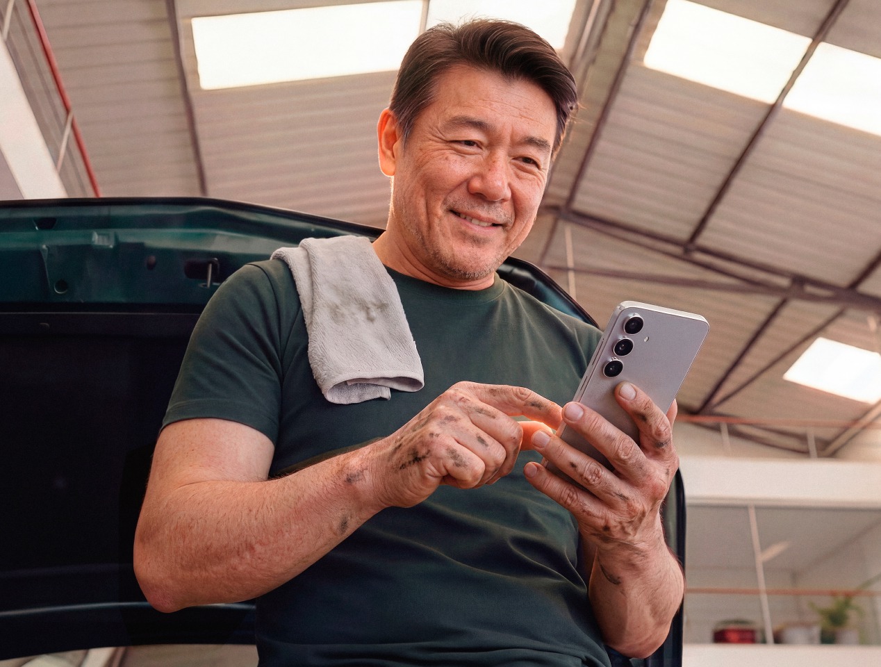 Mecânico em oficina sorrindo ao usar o celular, com pano apoiado no ombro