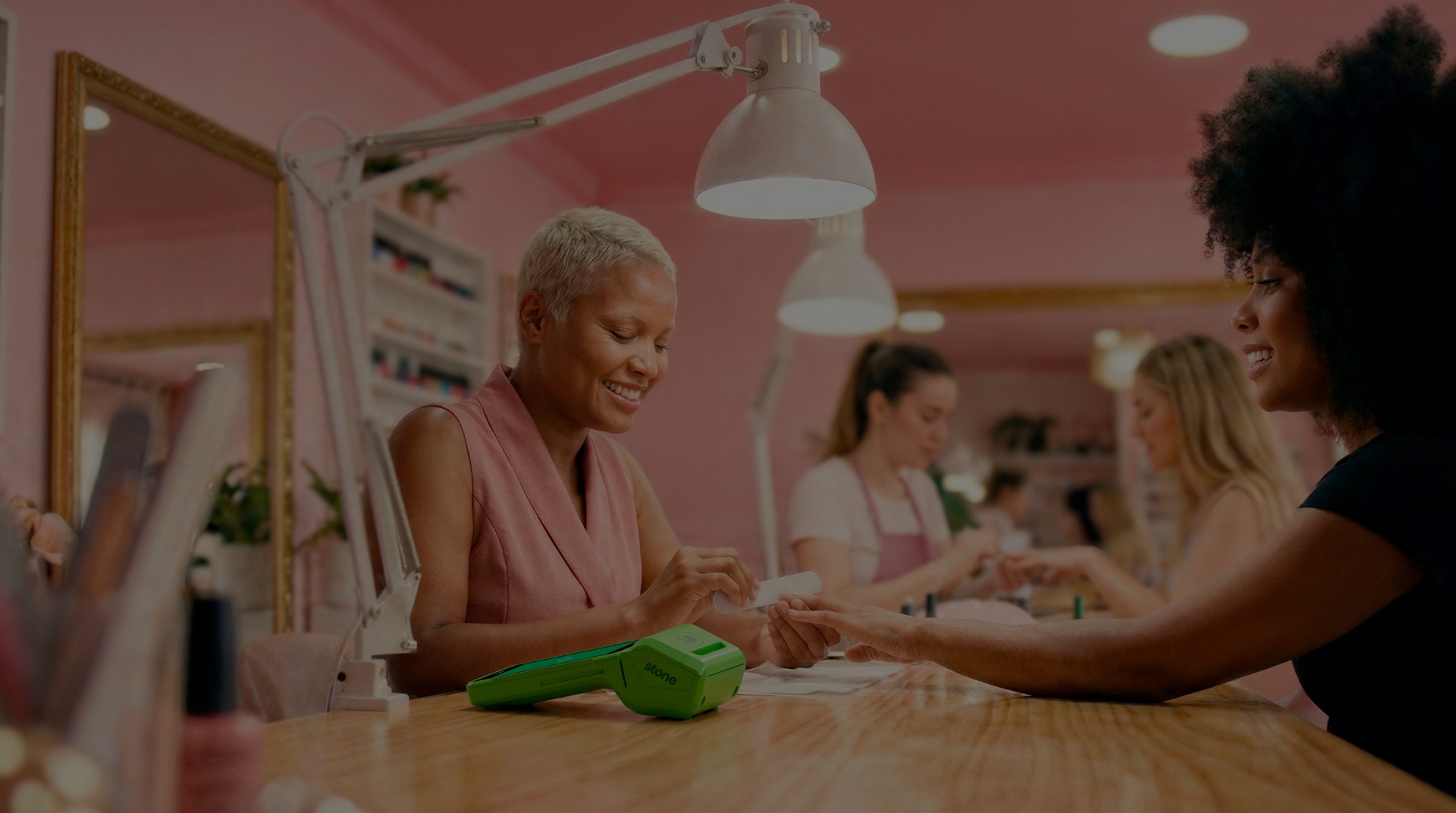 Manicure pintando as unhas de uma cliente em estúdio rosa, com a maquininha Stone ao lado