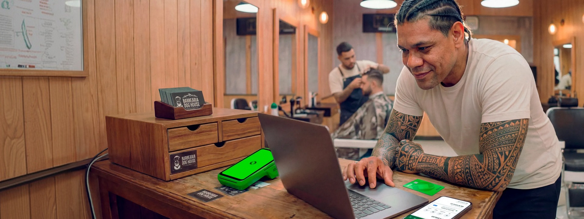Barbeiro tatuado em barbearia analisando vendas no notebook, com maquininha Stone e cartões da loja sobre a bancada