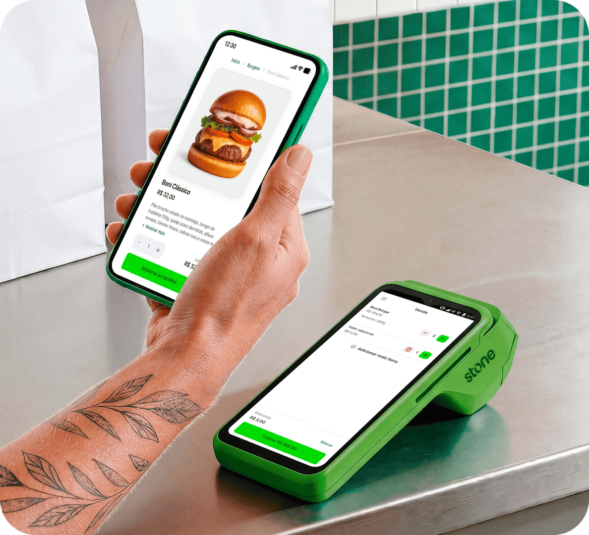 Empreendedora com uma camisa verde em seu restaurante e sentada no balcão de madeira, utilizando o seu notebook com uma mão e na outra mão o seu celular. Ao fundo alguns utensílios de cozinha.