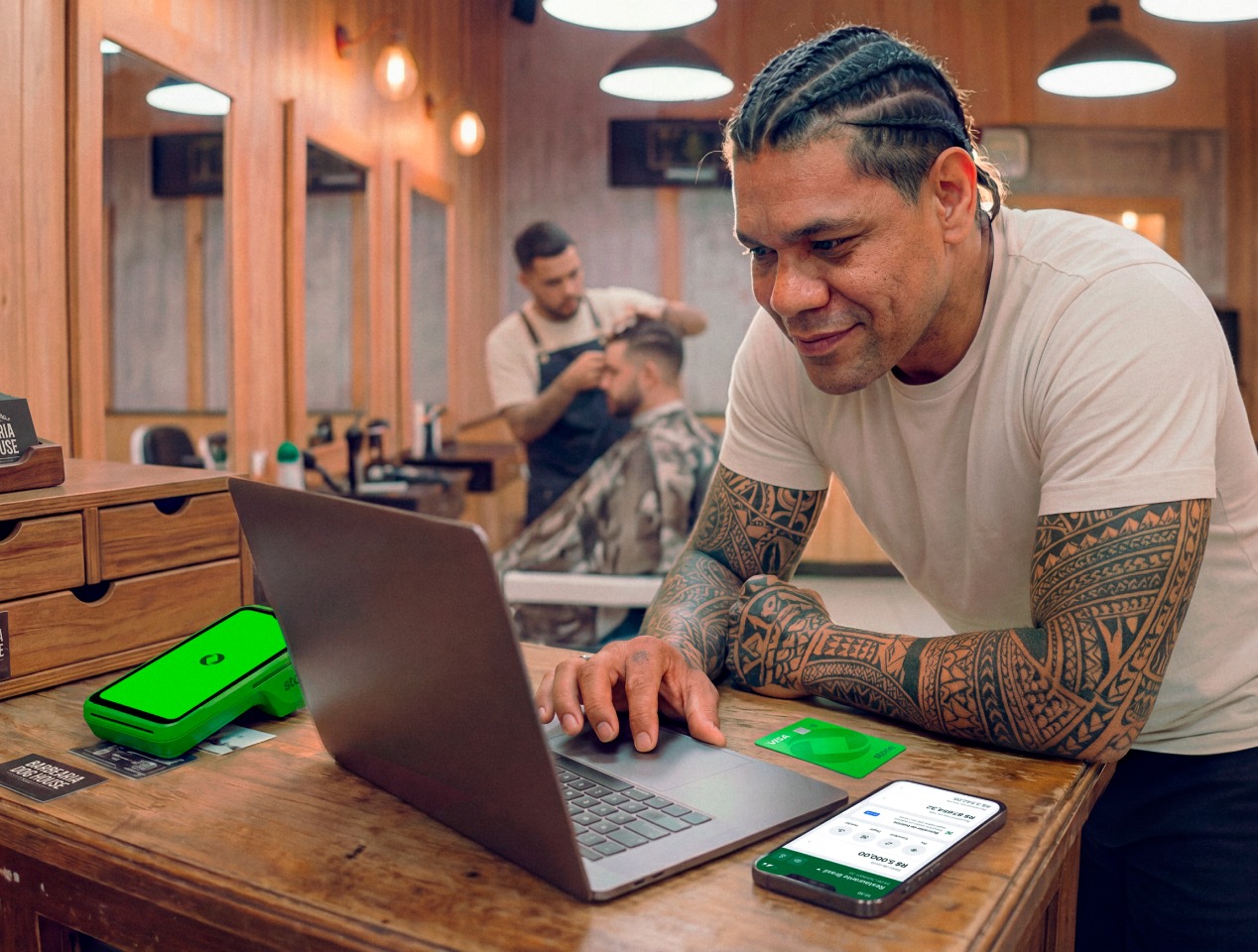 Barbeiro tatuado em barbearia analisando vendas no notebook, com maquininha Stone e cartões da loja sobre a bancada