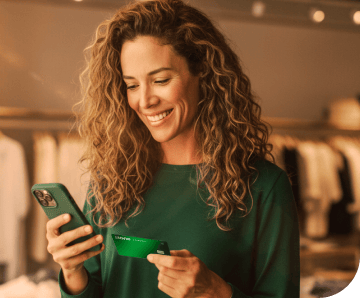 A empreendedora em sua loja de roupas, com linhas de costuras atrás está segurando um celular para enviar o link de pagamento aos seus clientes através do Messenger do Facebook, Whatsapp e Instagram.