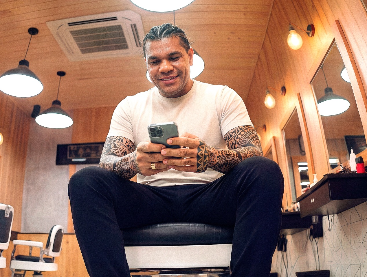 Barbeiro tatuado sentado em cadeira de barbearia consultando o celular