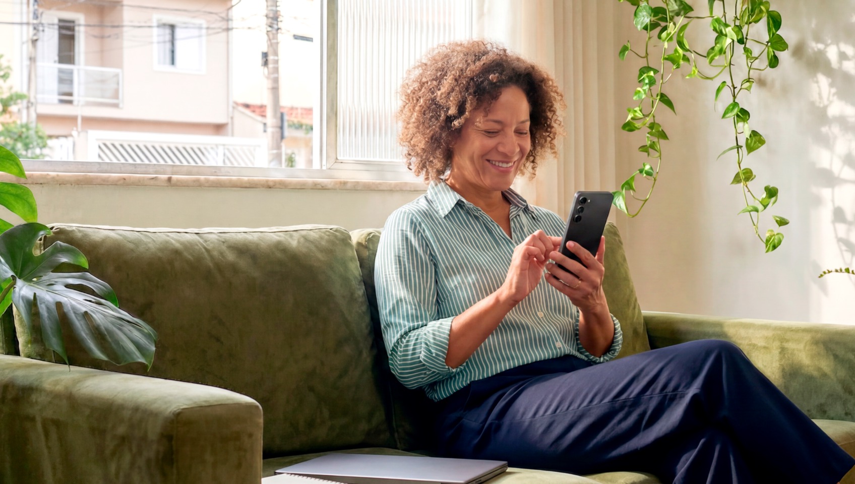 Mulher madura sorridente sentada no sofá de casa consultando o celular