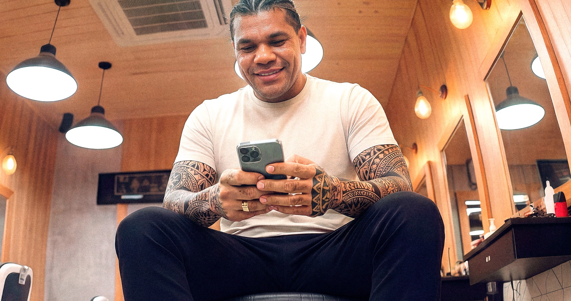 Barbeiro tatuado sentado em barbearia sorrindo enquanto consulta o celular