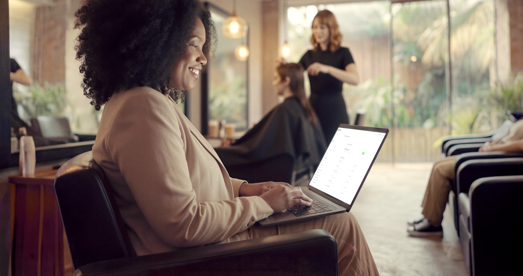 Empreendedora sorridente em salão de beleza usando o notebook enquanto outra cliente é atendida ao fundo