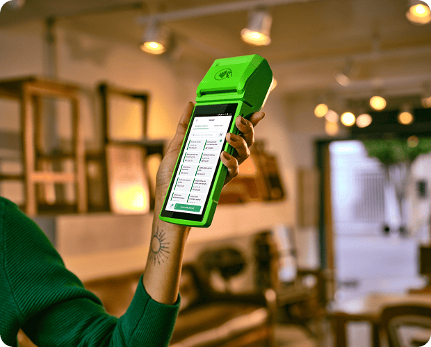 A empreendedora em sua loja de roupas, com linhas de costuras atrás está segurando um celular para enviar o link de pagamento aos seus clientes através do Messenger do Facebook, Whatsapp e Instagram.